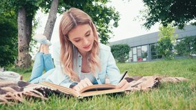 Student reading book and writing on notebook on plaid in park