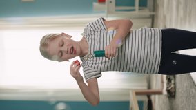 Funny blonde girl is having fun at home, blowing soap bubbles and smiling at camera sharing her positive emotions, entertaining indoor, concept of happy childhood, Vertical video, Slow motion.