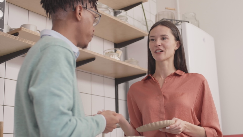Low angle view of attractive Caucasian brunette and young African man standing in the kitchen, having meal while communicating
