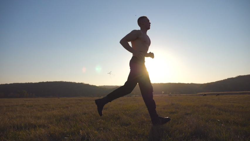 Young muscular man running through field with beautiful landscape at background. Male athlete trains in nature. Guy jogs at morning time. Unity with nature. Healthy active lifestyle. Side view Slow mo | Shutterstock HD Video #1056713684