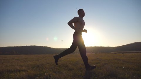 Young muscular man running through field with beautiful landscape at background. Male athlete trains in nature. Guy jogs at morning time. Unity with nature. Healthy active lifestyle. Side view Slow mo