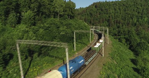 Freight train with cargo carriages an electric locomotive by two-sided Siberian railways near river in the forest mountains / Aerial drone mid view at summer sunset