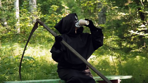 Grim Reaper with Scythe Drinking Coffee while Sitting on a Bench in a Park. Slow Motion. Life and Death, Another World and Halloween Concept