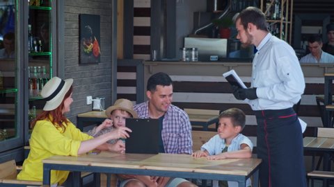 lifestyle, charming married couple with young male children enjoying a family vacation and ordering waiter food and drinks from cafe or restaurant menu