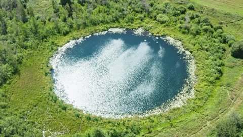 Aerial view camera locked over lake of perfectly round shape. Sky and sun light reflected in clear turquoise water of pond surrounded by trees and plants. Ripple on water surface, windy summer day