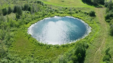 Aerial view drone flies over lake of round shape. Cloudy sky and sun light reflected in clear turquoise water of pond surrounded by trees and plants. Ripple on water surface, windy sunny summer day
