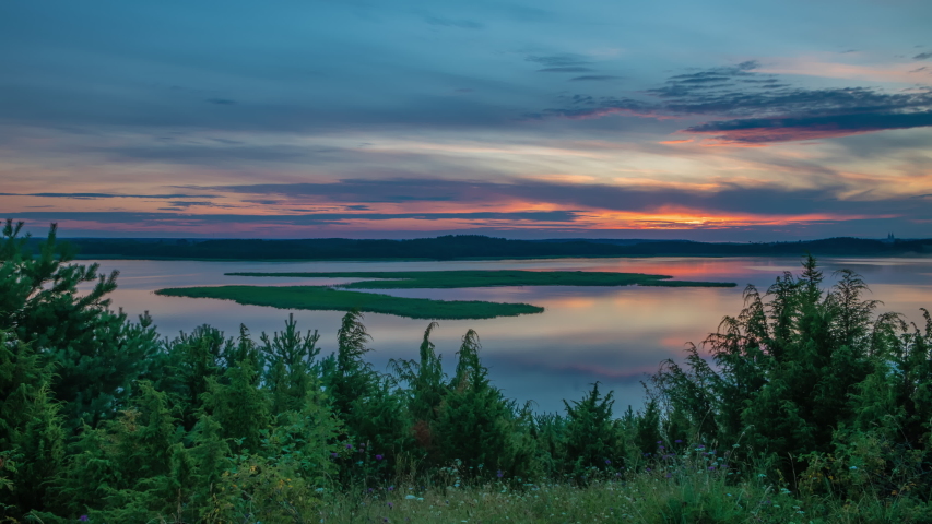 Time-lapse sunrise over lake in national park. Water surface in motion. Multicolored sky moving clouds. Green pine trees in front of camera, forest on horizon. Bright sun rises over colorful skyline