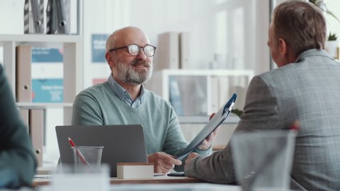 Senior male executive manager greeting colleague with handshake, checking documents in file, smiling and talking with man during office workday