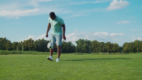 Handsome african man in casual clothes bouncing soccer ball on legs on green grass in public park. Active black guy practicing football, demonstrating skills of controlling and juggling ball in nature
