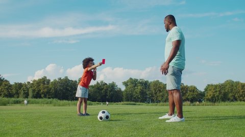 Adorable preschooler mixed race football referee with curly hair blowing whistle, showing red card to african father on green field while family playing soccer game during weekend in summer nature.
