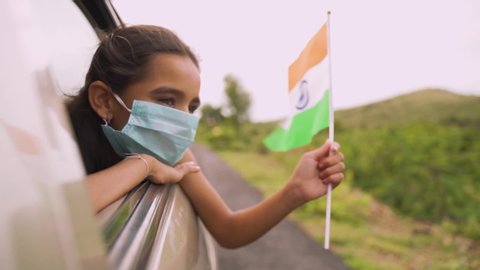 Young girl kid with a medical mask holding an Indian flag in moving car window. Concept of celebrating Independence or republic day during coronavirus or covid-19 pandemic.