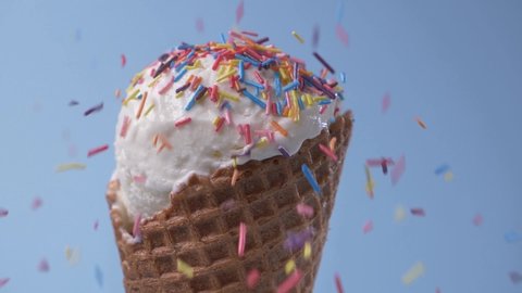 Rotating vanilla white ice cream in waffle cone covered with rainbow sugar sprinkles topping on blue background. Sweet dessert. Delicacy.