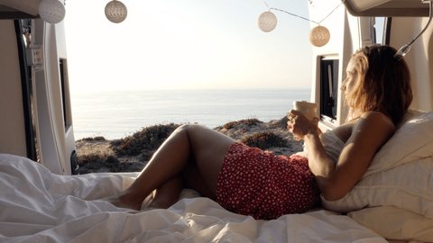 Woman drinking from cup in camper van enjoys stunning coastal views from her bed. Young hipster woman living the dream in her camper van. Digital nomad freedom concept 