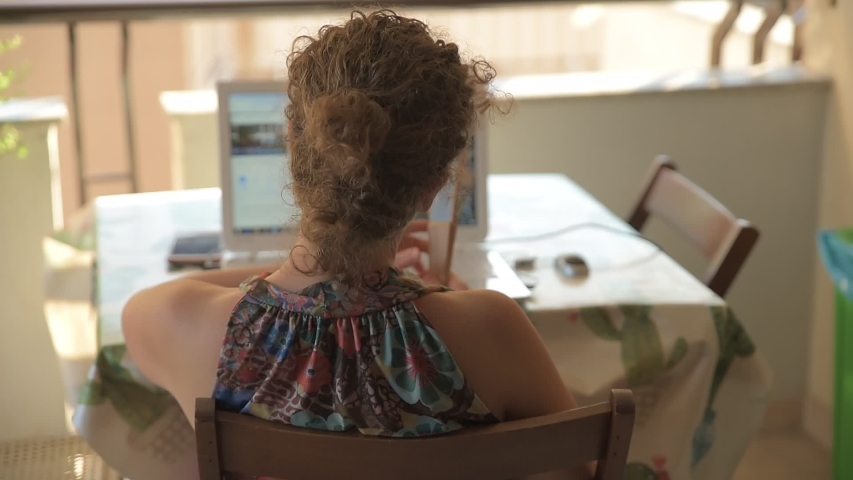 Young Caucasian woman shooting from the back using her laptop sitting at the table outside on the balcony. Woman blowing with her fan, works from home outside on the balcony.