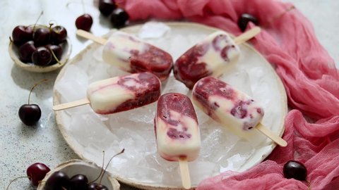 Fresh cream and cherry homemade popsicles placed on white ceramic plate with fruits and textile. Top view, flat lay.