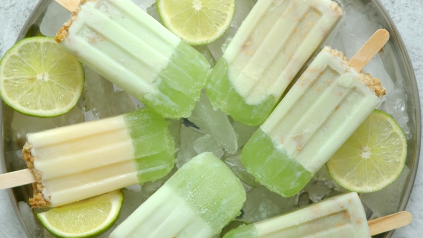 Summer refreshing homemade lime popsicles placed on metal tray with chipped ice over stone background, top view. Flat lay | Shutterstock HD Video #1056737933