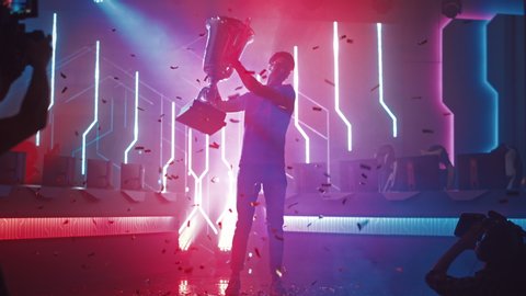 Professional Video Games Player Tournament Winner Celebrates Victory Cheering and Holding Trophy. Big Stylish Neon Bright Championship Arena doing Pro Computer Gaming Event with Online Streaming