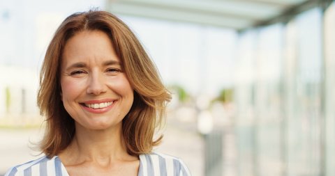 Portrait shot of beautiful Caucasian woman with fair hair looking at side, turning face to camera and smiling outdoors on sunny summer day. Happy female at street. Close up of cheerful lady.