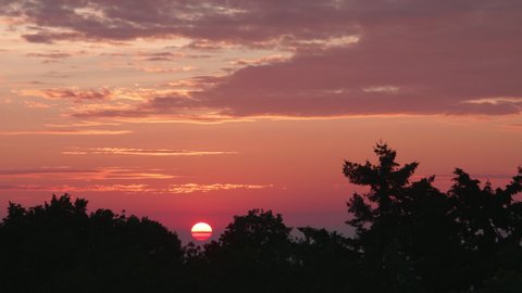 time lapse – red sun rising slowly in the morning dawn over tree silhouettes with red clouds in the sky