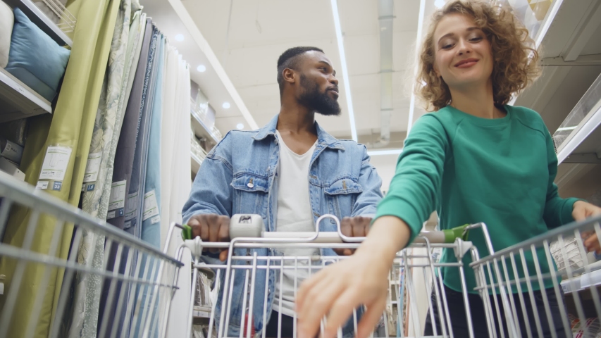 Happy multiethnic couple buying pillows in house improvement store. Low angle view of cheerful husband and wife with shopping cart purchasing decorations for home in furniture store Royalty-Free Stock Footage #1056738971