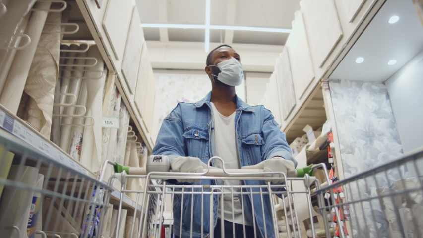 African man wearing protection facemask choosing wallpaper in hardware store. Low angle view of afro guy pushing shopping cart buying materials in house improvement store wear safety mask and gloves Royalty-Free Stock Footage #1056739022