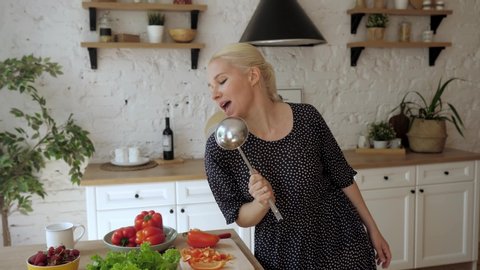 Smiling slim young woman holds knife cuts cherry tomatoes preparing salad cooking vegetable dish moves to music listens song dancing in kitchen feels carefree. Merrily dancing and singing in the ladle