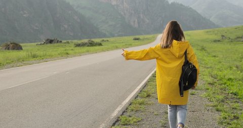 Young woman traveller hitchhiker in yellow raincoat with backpack is on an asphalt road trying to vote thumb up, stop the car. Traveling in natural park with huge mountains. Vacation concept.: stockvideo