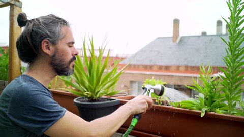 Young bearded man watering flowers on urban garden. Slow motion
