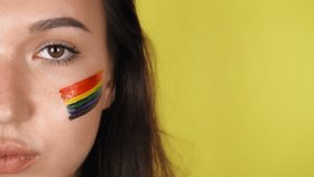 Woman with a pattern of a rainbow on her face close-up. The LGBT flag is painted on the face. Yellow background. High quality 4k footage