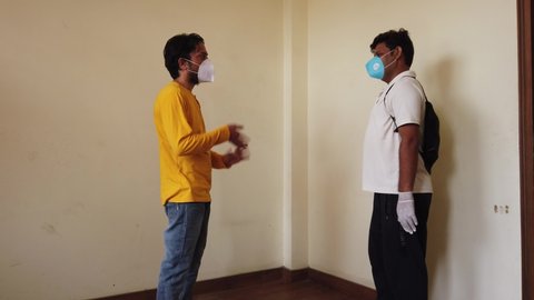 Two male friends in a contactless greetings each other indoor wearing mask being careful passing each other during coronavirus pandemic in new normal post global pandemic due to safety concerns 