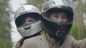 Smiling happy bikers in helmets shooting selfie video while riding on motorcycle. Portrait of Caucasian man and woman driving scooter on cloudy day outdoors. Camera zooming out.