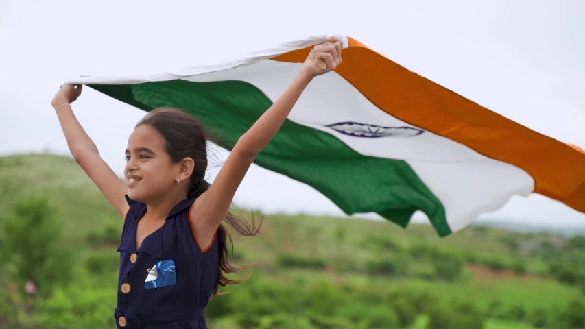Smiling girl Kid holding waving Indian flag in the air on top of hill - Concept of patriotism, celebration Independence or republic day.