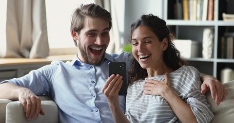 Surprised young family couple looking at mobile screen, feeling excited about online lottery win message. Overjoyed spouses winning giveaway in social networks, celebrating success together at home.