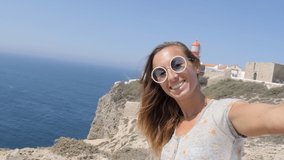 Woman travelling in Sarges Portugal, takes a selfie with lighthouse in the background. Young female selfie with coastal views 
