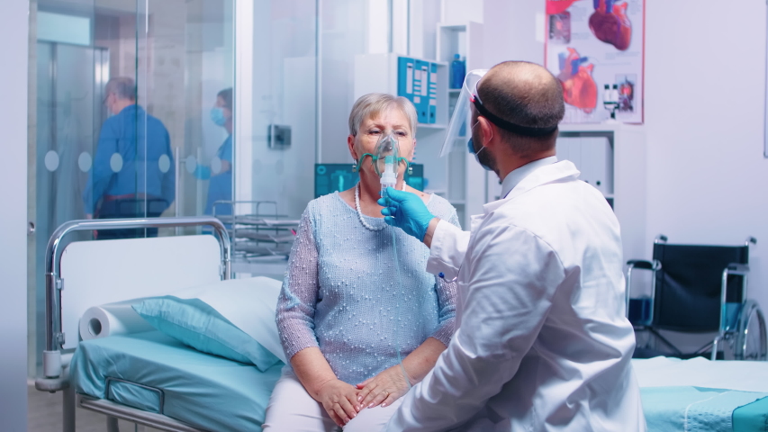 Doctor putting an oxygen mask on old retired senior woman during COVID-19 coronavirus outbreak in modern private hospital or clinic. Fighting infection and disease medicine and quarantine | Shutterstock HD Video #1056785636