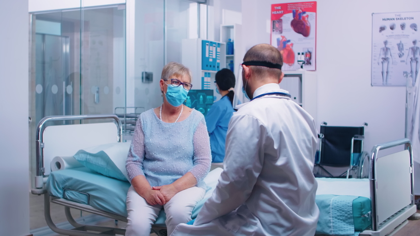 Medical stuff in visor explainingX Ray result to old retired woman sitting on hospital bed during COVID-19 pandemic. Nurse checking with old man in background. Modern private clinic or healthcare | Shutterstock HD Video #1056785690