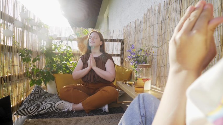 tranquil young woman meditates in lotus position with friend holding small crystals in palms on decorated terrace at home on sunny summer day