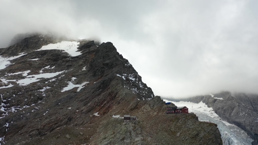 Drone shot of the mountain peak next to Dossen Hut in Switzerland, with dramatic and moody clouds coming over the snow covered top. Massive glacier in the background in the valley.