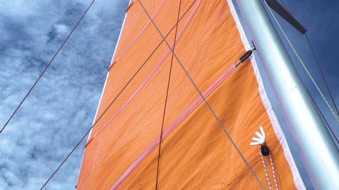 View of orange mainsail of yacht against the blue sky on summer. Stainless mast of a sailboat with attached ropes and mainsail. Slow motion shot.