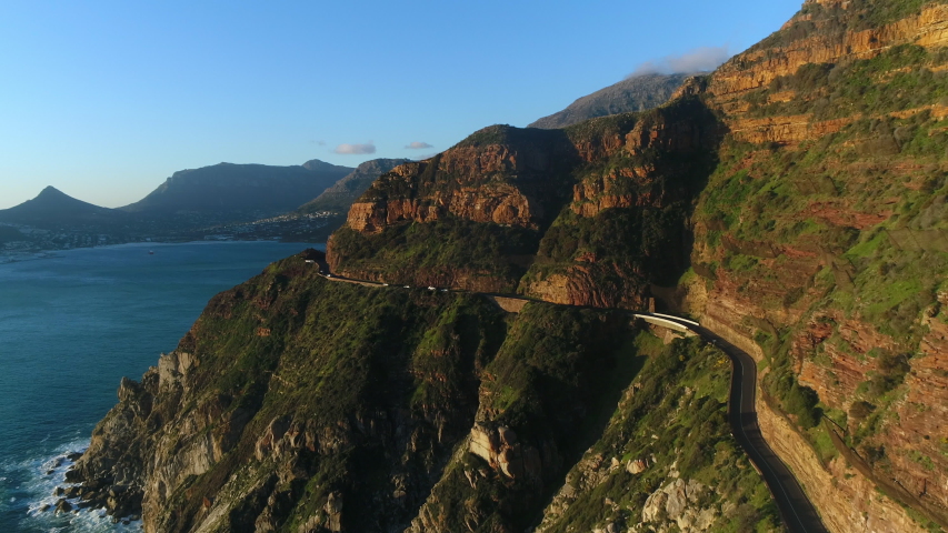 Aerial view of cars driving along Chapman's peak mountain pass while the sun is setting. Camera tracks forward to he road and slowly ascends over one of the peaks. Royalty-Free Stock Footage #1056798167