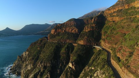 Aerial view of cars driving along Chapman's peak mountain pass while the sun is setting. Camera tracks forward to he road and slowly ascends over one of the peaks.
