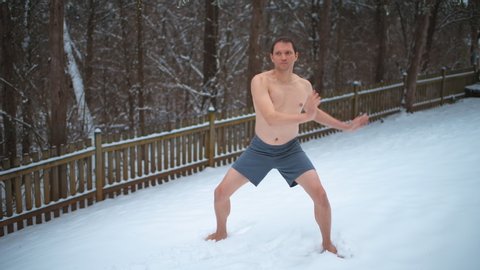 Shirtless man doing cold exposure therapy standing horse stance barefoot yoga pose in house backyard with snow snowing blizzard white winter storm snowflakes weather