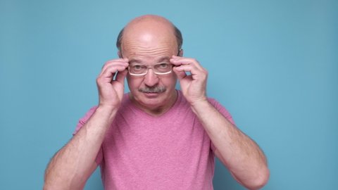 Senior hispanic man looking through glasses being shocked. Studio shot on blue wall.