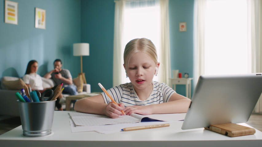 Small girl is sitting at table at home, studying online, writing something in her notebook, using tablet computer, gadget, parents are busy with their affairs, Foreground, Slow motion.