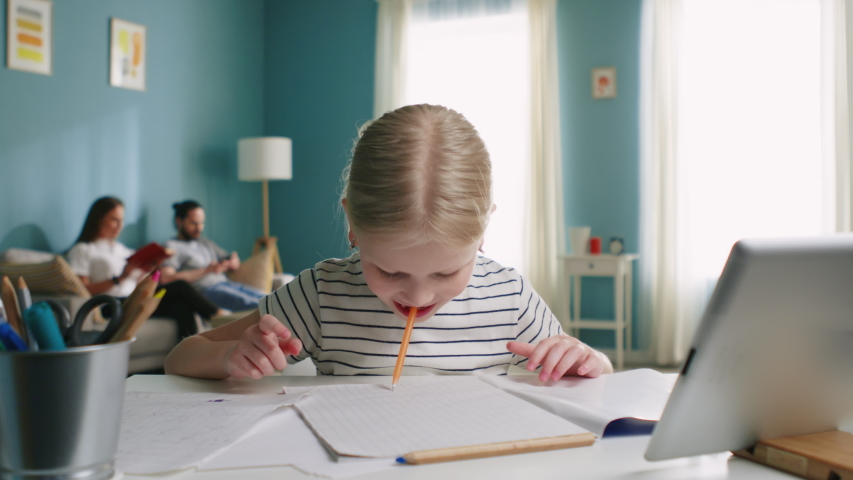 Funny girl is sitting at desk at home, and has fun instead of studying, drawing by her mouth, enjoys her childhood, parents are on background, Slow motion.