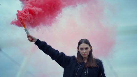 Portrait of young woman standing on street with smoke bomb in hand, ambitious woman holding smoke grenade at protest, closeup serious activist looking at camera on street