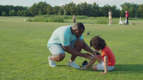Caring loving black father with antiseptic and wet wipe providing first aid, treating and cleansing wound on sons scraped knee after fall while preschool mixed race child sitting on green field