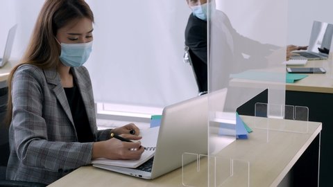 Normal view, medium shot of asian male and female employee talk to each other with facial mask on, while working on own desk seperated away to keep in distance with clear partition on the table.