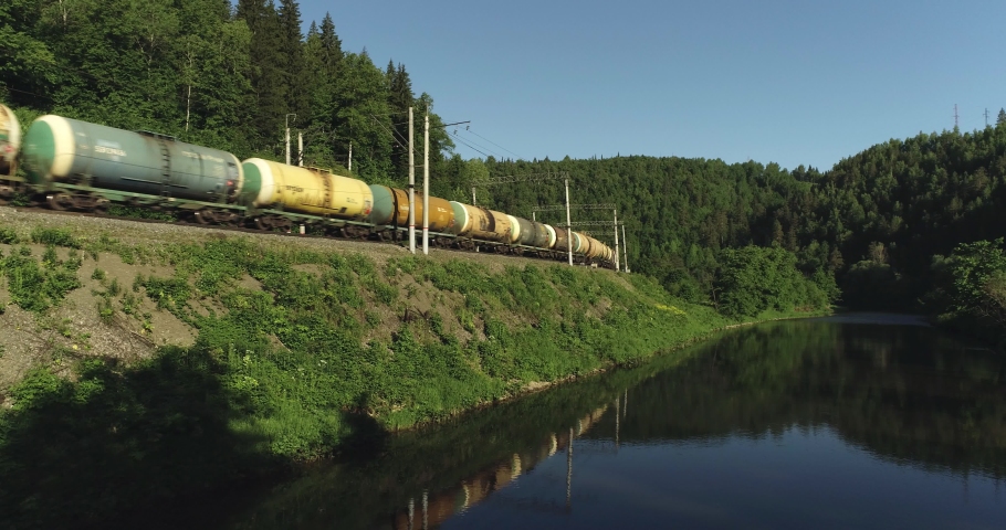 Freight train with with oil tank carriages an electric locomotive by two-sided Siberian railways near river in the forest mountains / Aerial drone mid view at summer sunset Royalty-Free Stock Footage #1056813557
