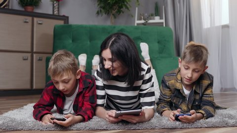 Family concept where attractive contented cheerful mother and her two 8-10s sons which lying on fluffy carpet and revisioning modern apps on tablet pc 4k
